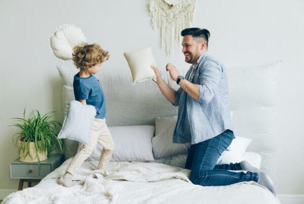Young man fighting pillows with little son having fun on bed in apartment