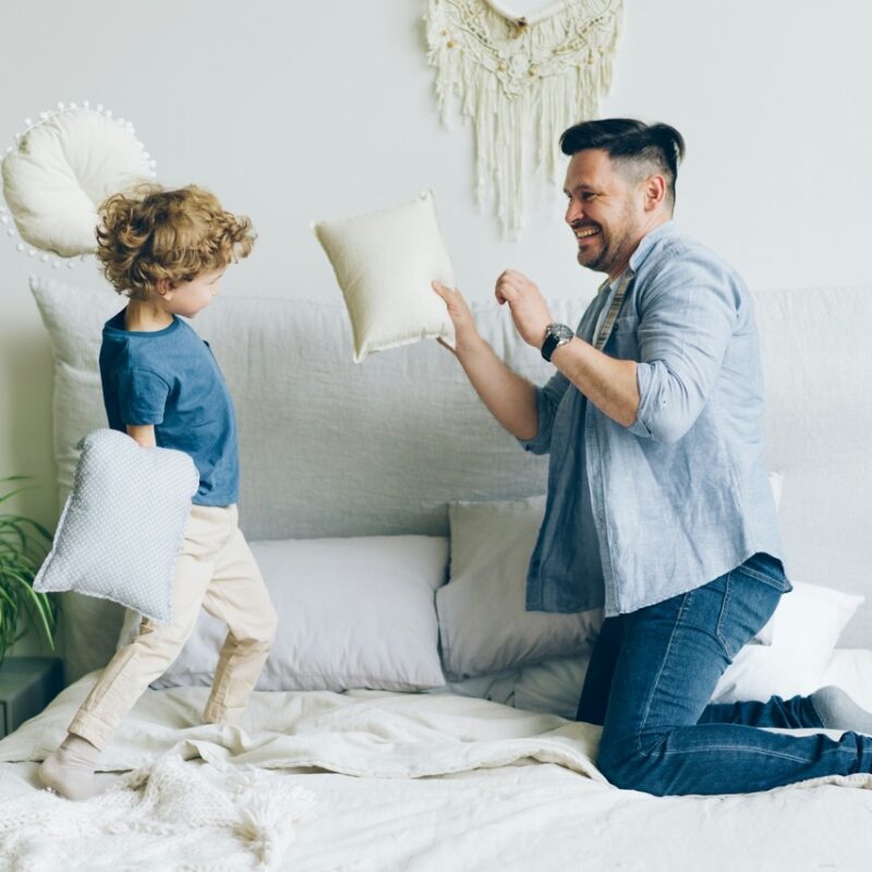 Young man fighting pillows with little son having fun on bed in apartment